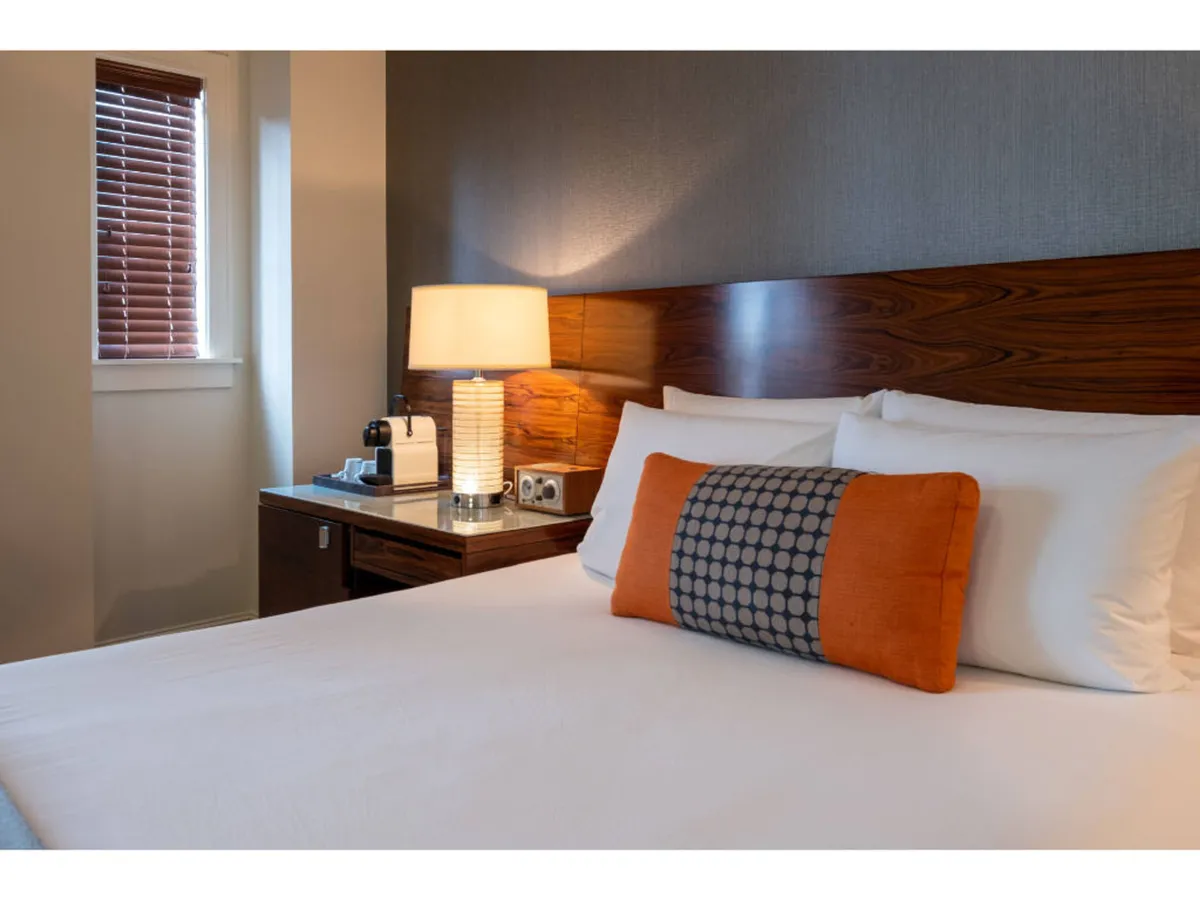This interior image features a cozy, inviting king bed adorned with plush decorative pillows, flanked by a stylish nightstand and a softly glowing table lamp. Natural morning light filters gently into the room, creating a serene, restful ambiance ideal for relaxation or enjoying coffee by the window.
