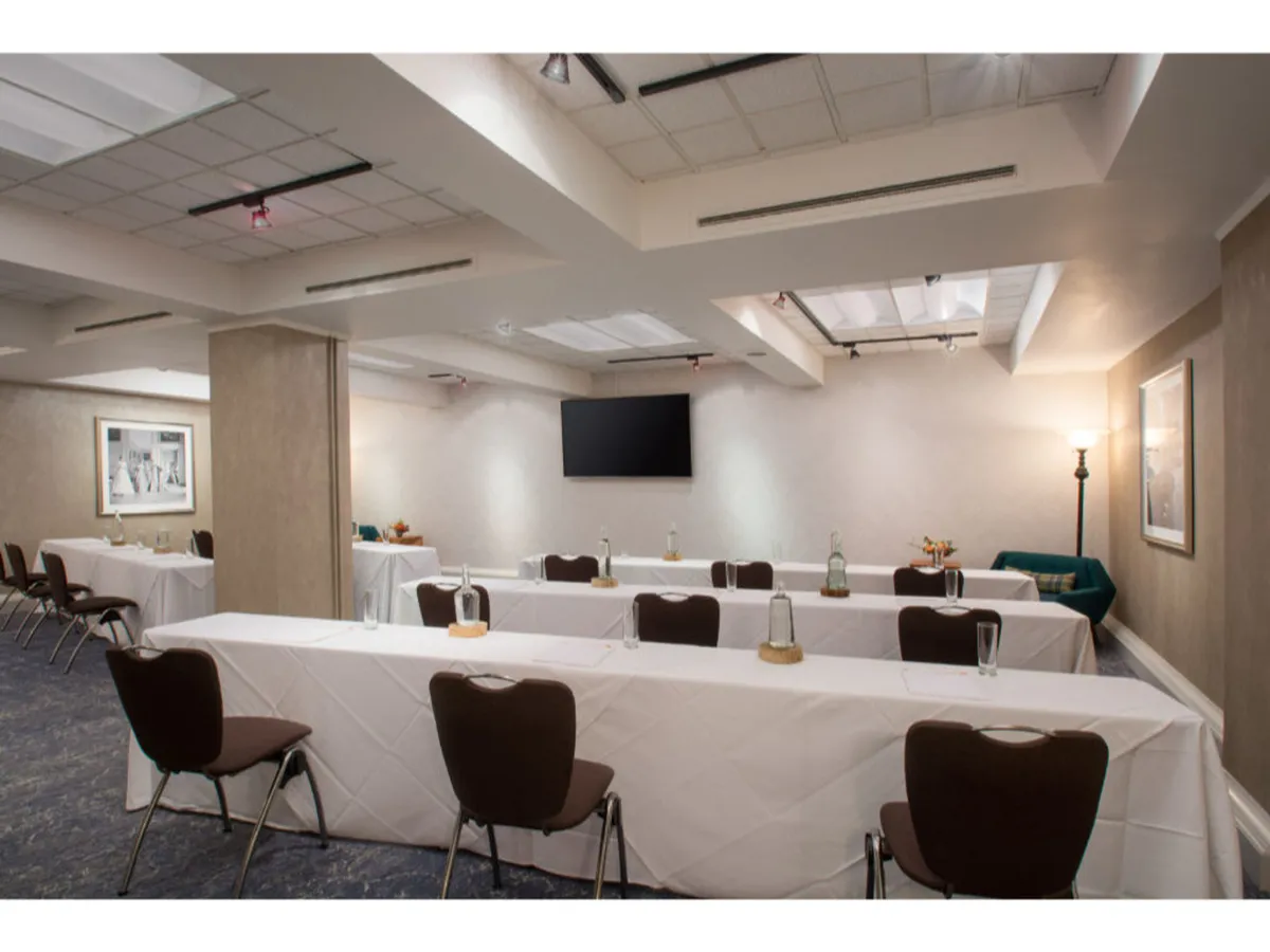A professional meeting room setup with long white tablecloths, neatly arranged chairs around tables, a TV screen for presentations, and table decorations including water bottles and glasses. The environment looks conducive for team meetings and workshops.