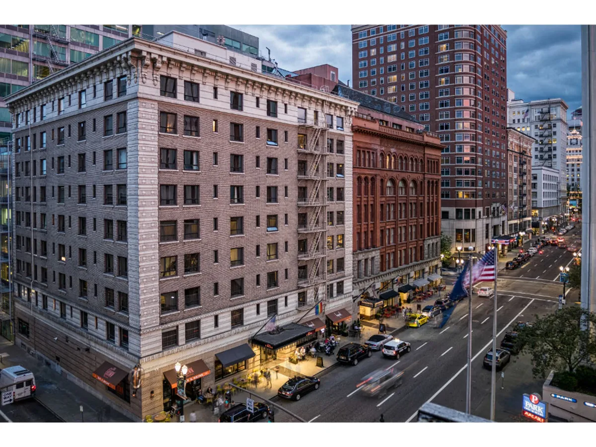 A dynamic city street view shows a historic red brick building alongside a modern skyscraper, lined with cafes, bustling pedestrians, and American flags. The street scene encapsulates the architectural diversity and lively atmosphere of the Pearl District and downtown Portland.