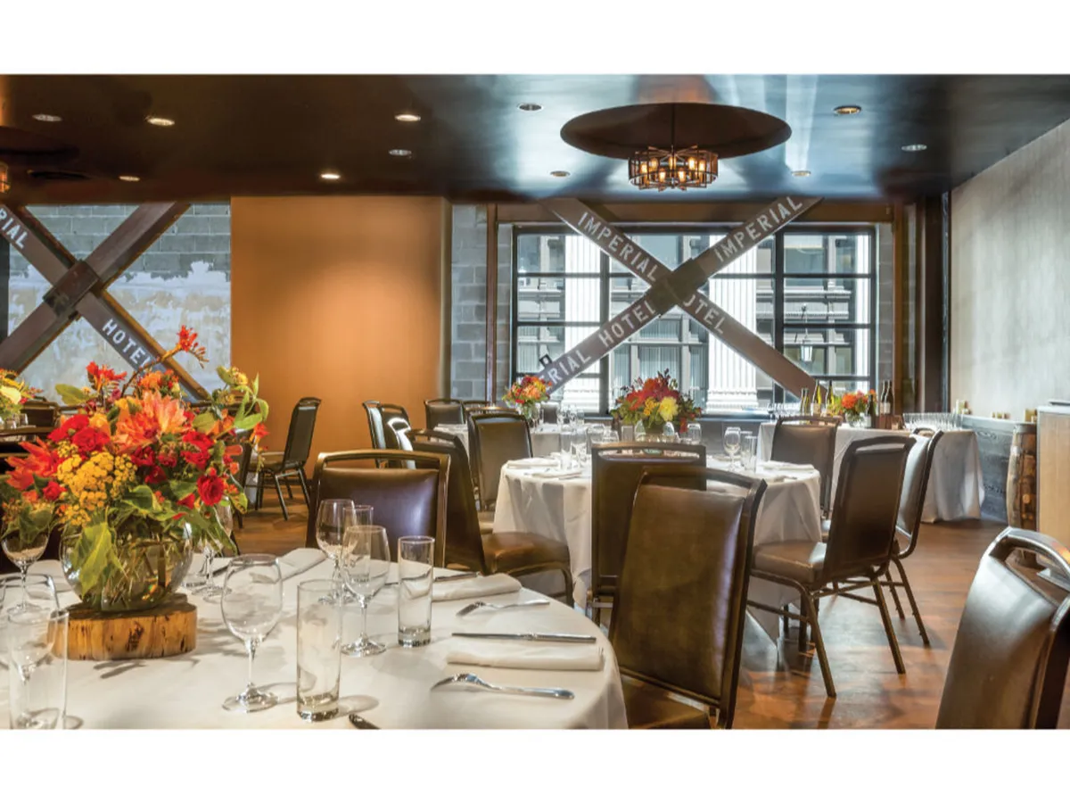 A dining area set with floral centerpieces, tables ready for guests, and large windows that offer sweeping views of the city beyond. Sunlight illuminates the space, creating a lively and elegant atmosphere ideal for gatherings and moments of enjoyment.