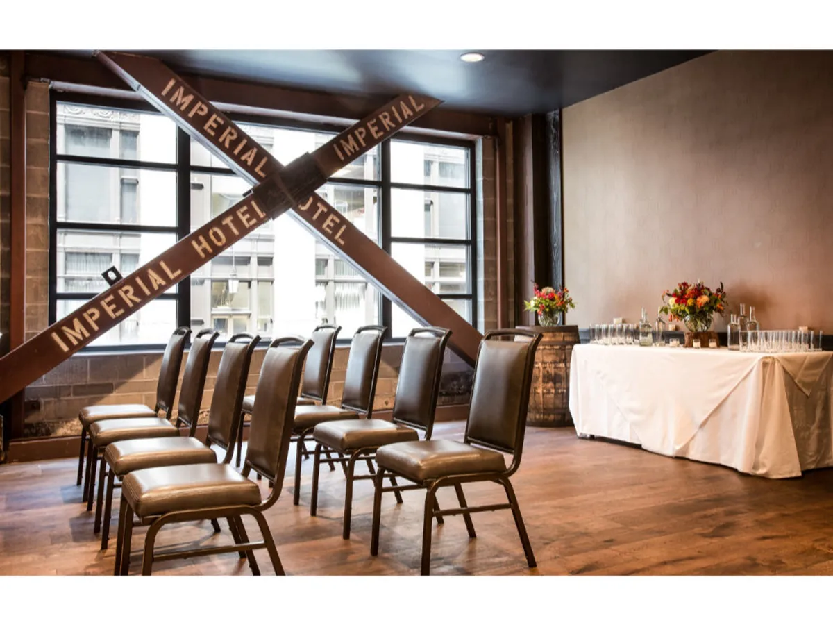 A sophisticated interior event space featuring rows of chairs, a decorative crossbeam, a central floral arrangement, and a serving table. The setting is arranged for gatherings and can accommodate meetings, conferences, or social events in a refined atmosphere.