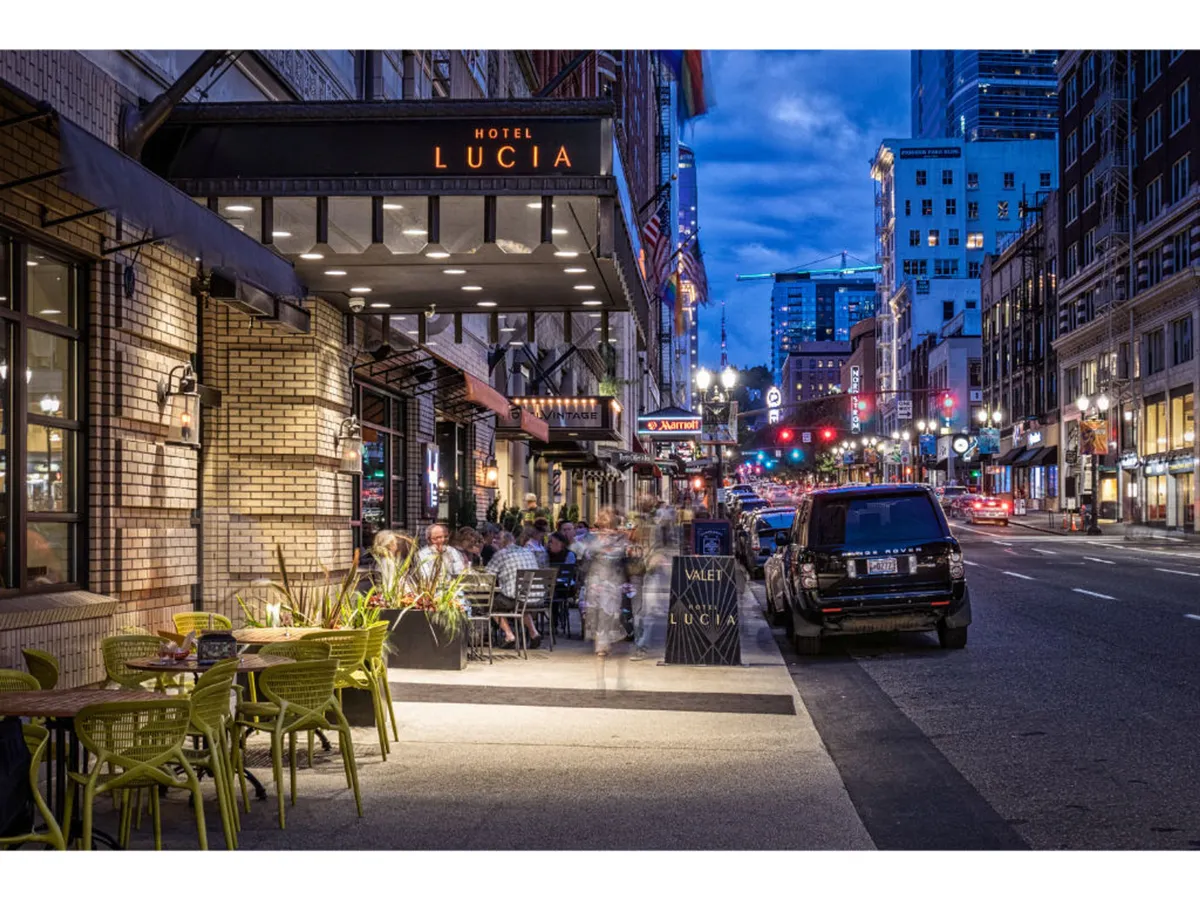 A bustling exterior scene featuring the Hotel Lucia sign, outdoor seating, and city street ambiance. The evening lighting and visible activity evoke the energetic, social atmosphere of Portland’s downtown arts district, ideal for launching a culture walk.