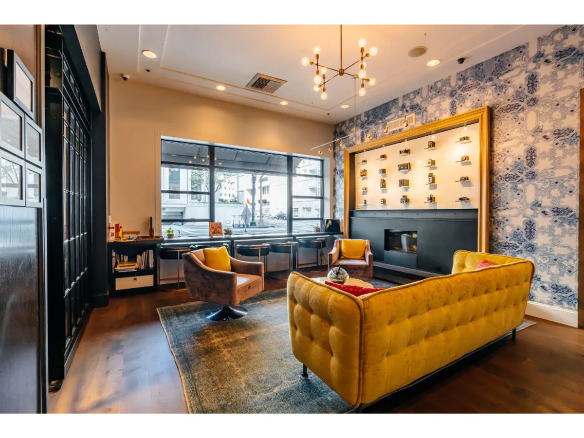 The image features a stylish, comfortable lounge area with a yellow tufted sofa, chandeliers, blue patterned wallpaper, and large windows letting in natural light. The space is designed for social gatherings, reading, or relaxing, creating an inviting atmosphere for guests to unwind or connect with others.