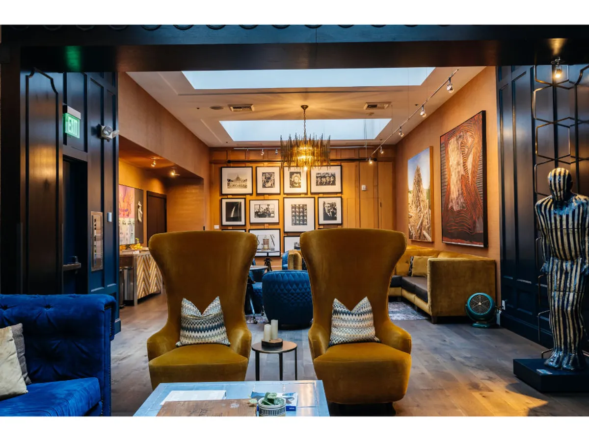 This image features a cozy lounge area with two large yellow armchairs, a blue couch, a modern chandelier, and various framed photographs on the wall. A sculptural figure accentuates the artistic atmosphere, and the space feels contemporary yet welcoming.