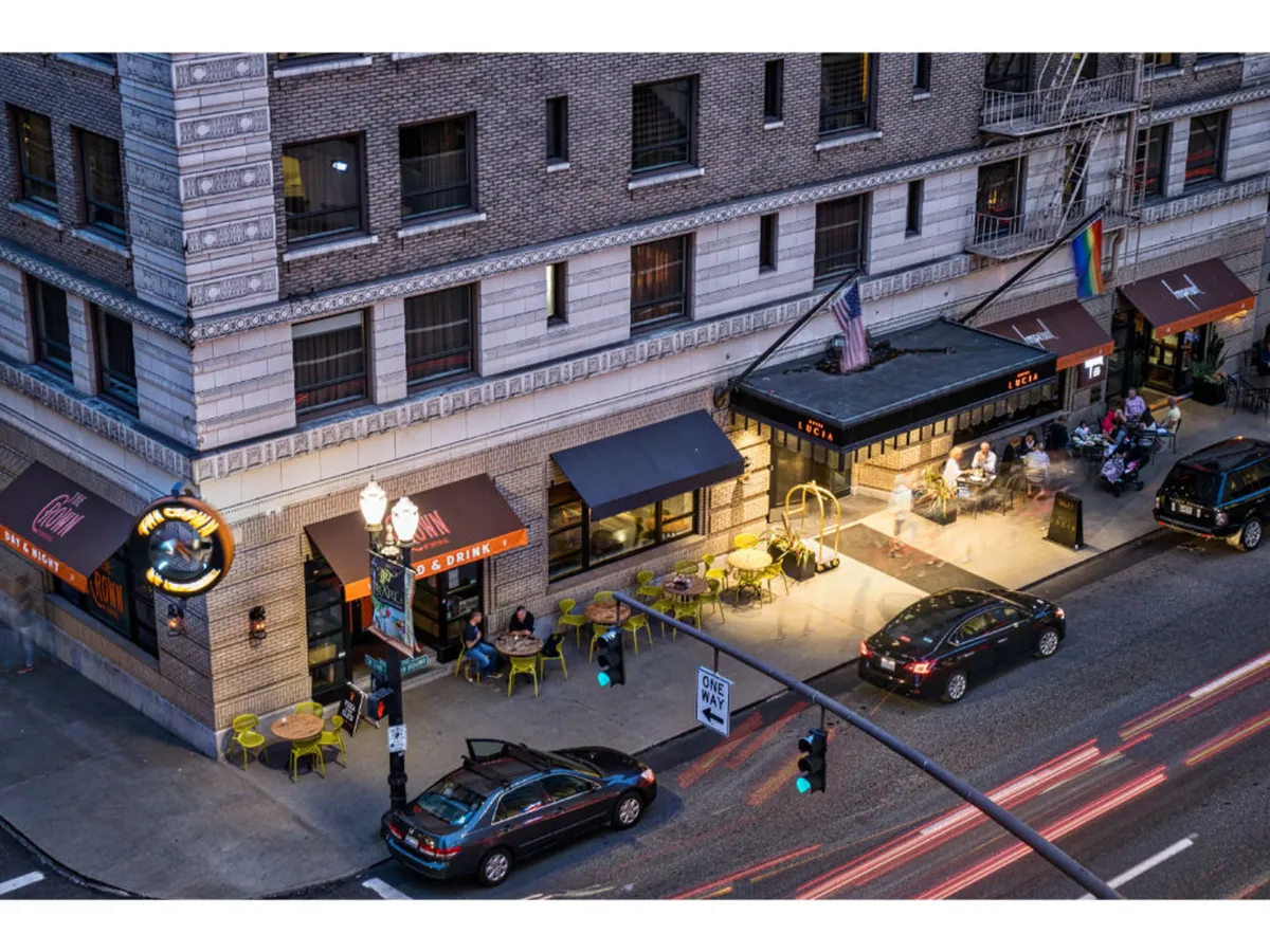 An animated exterior scene showcasing a bustling outdoor seating area at a cafe, illuminated by a vibrant neon sign. Groups of people are dining al fresco, enjoying drinks and conversations. Cars pass by on the street, adding to the urban nightlife ambiance.