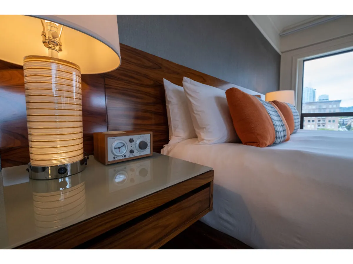 This image captures a serene moment in the bedroom, focusing on the bed with decorative pillows, a nightstand with a table lamp, and soft natural light. The scene evokes relaxation—perfect for reading, resting, or enjoying a quiet evening with music.