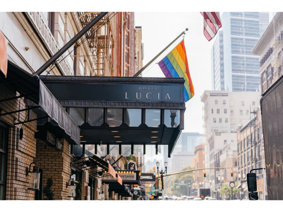 This exterior shot captures the Hotel Lucia's sign flanked by a rainbow flag and an American flag, set against the urban streetscape. The welcoming street scene suggests easy access to local art venues and cultural hotspots, reinforcing the hotel's connection to Portland's vibrant community.